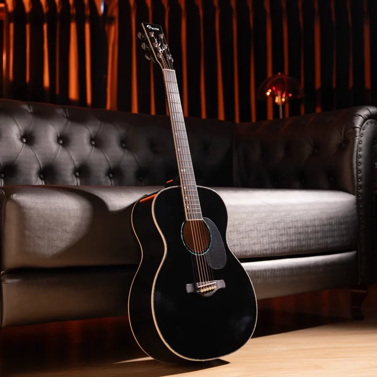 Black acoustic guitar leaning against a dark tufted leather sofa in a dimly lit room, on a wooden floor. ขายราคาพิเศษ