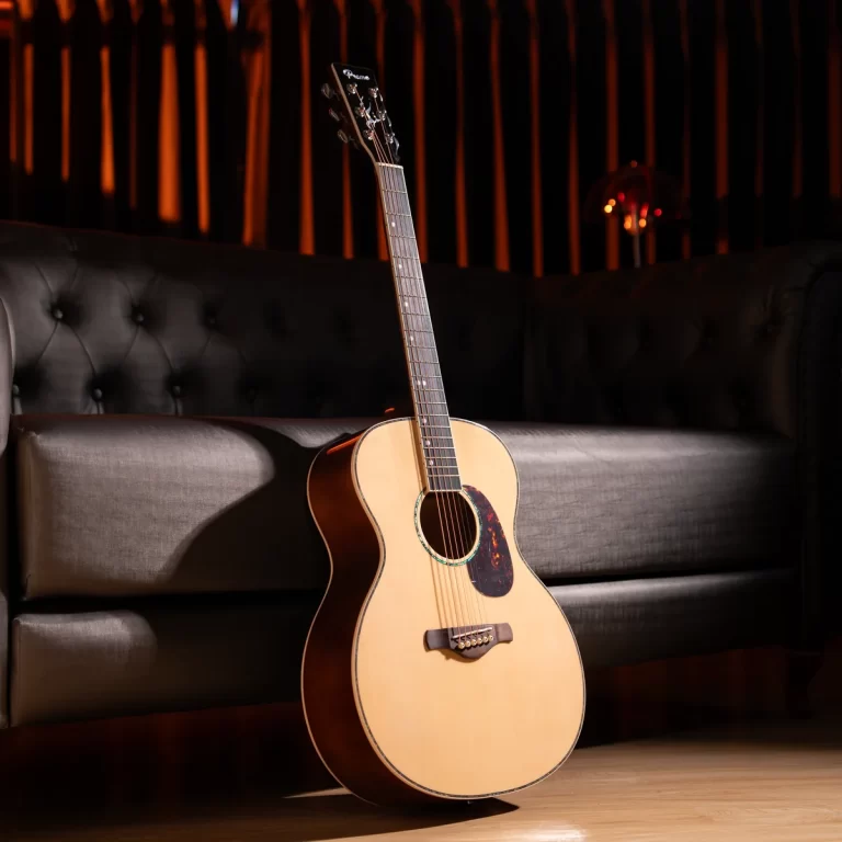 Acoustic guitar resting on a wooden floor beside a black tufted sofa in a moody lounge with warm orange lighting. ขายราคาพิเศษ