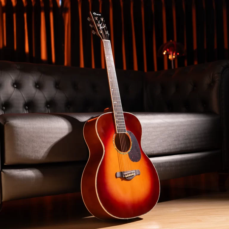Sunburst acoustic guitar resting upright on a light wood floor beside a black tufted sofa, in a warm-lit room. ขายราคาพิเศษ
