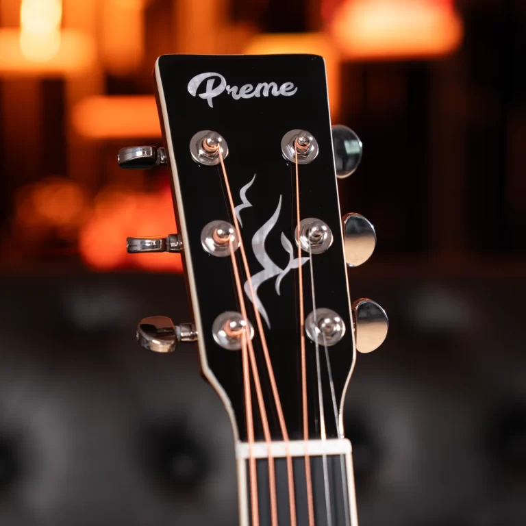 Close-up of a black guitar headstock with six chrome tuners, copper strings, and a white flame inlay; brand name 'Preme' visible on the headstock, warm blurred background. ขายราคาพิเศษ