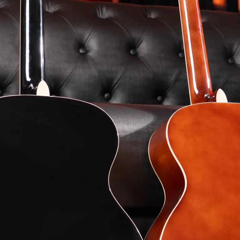 Two acoustic guitars lean against a black tufted leather sofa: a black guitar on the left and a sunburst guitar on the right, necks visible. ขายราคาพิเศษ