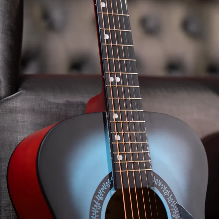 Close-up of a blue sunburst acoustic guitar neck with frets and white fret markers resting against a gray sofa. ขายราคาพิเศษ