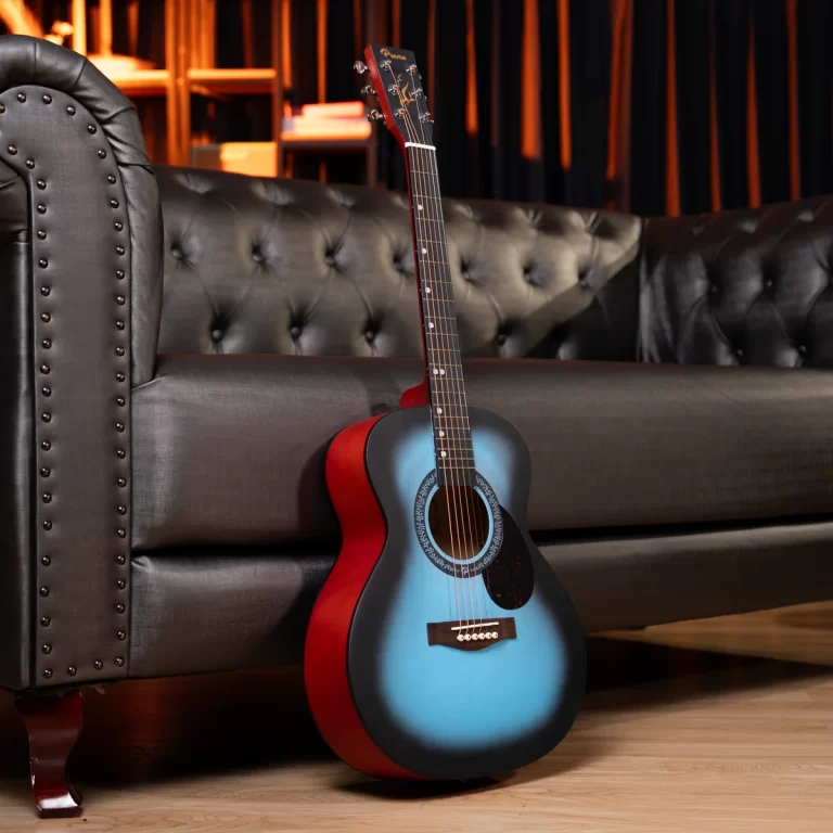 Blue-to-navy gradient acoustic guitar with red sides resting against a tufted black sofa in a cozy living room/studio. ขายราคาพิเศษ