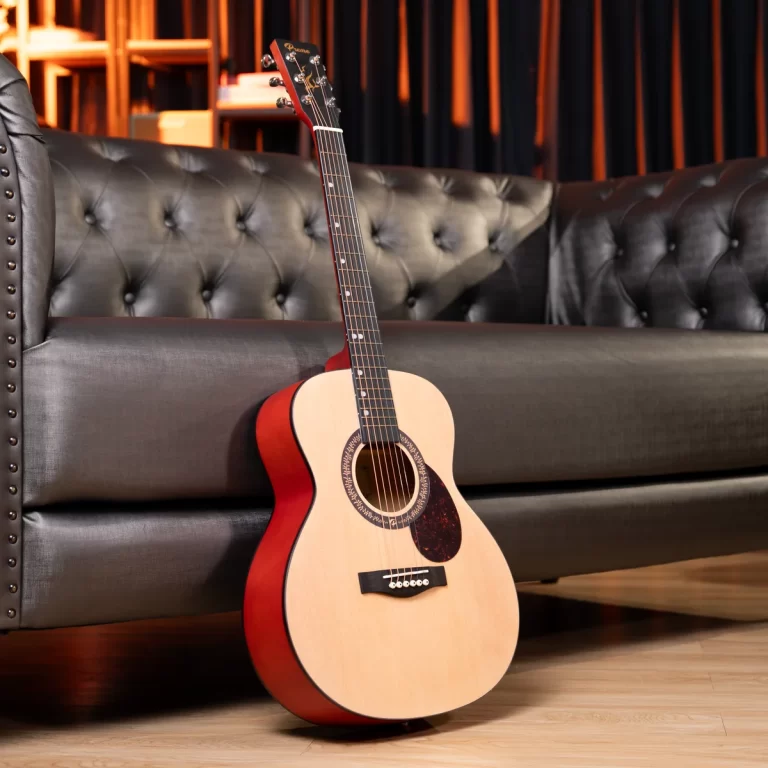Acoustic guitar with natural top and red sides resting on a wooden floor beside a tufted black leather sofa in a cozy living room setting. ขายราคาพิเศษ