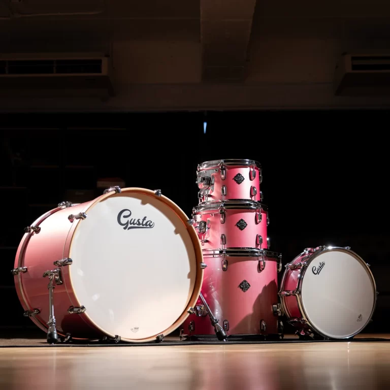 Pink drum kit arranged on a dim stage, featuring a large bass drum with the Gusta logo and stacked toms with chrome hardware. ขายราคาพิเศษ