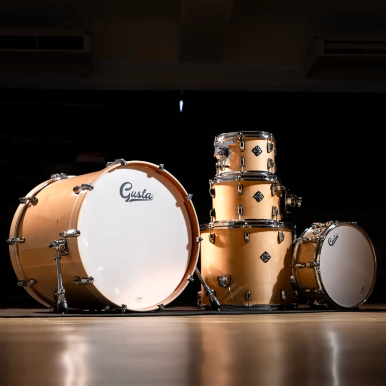 Peach-colored drum kit on a dark stage, featuring a large bass drum with the Gusta logo and stacked toms nearby. ขายราคาพิเศษ