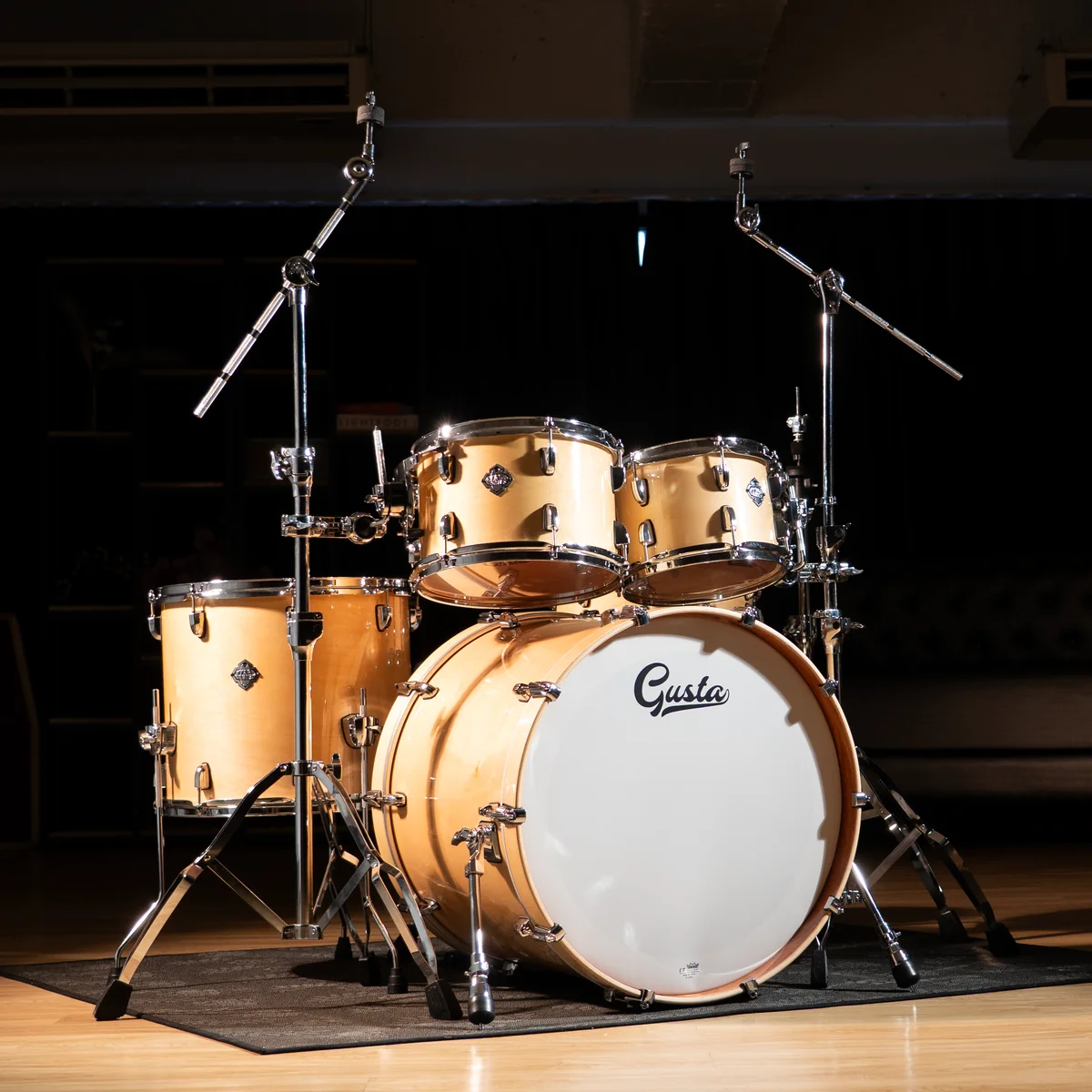 Beige drum set with a white bass drum head labeled 'Gusta' on a dim stage, ready for a performance.