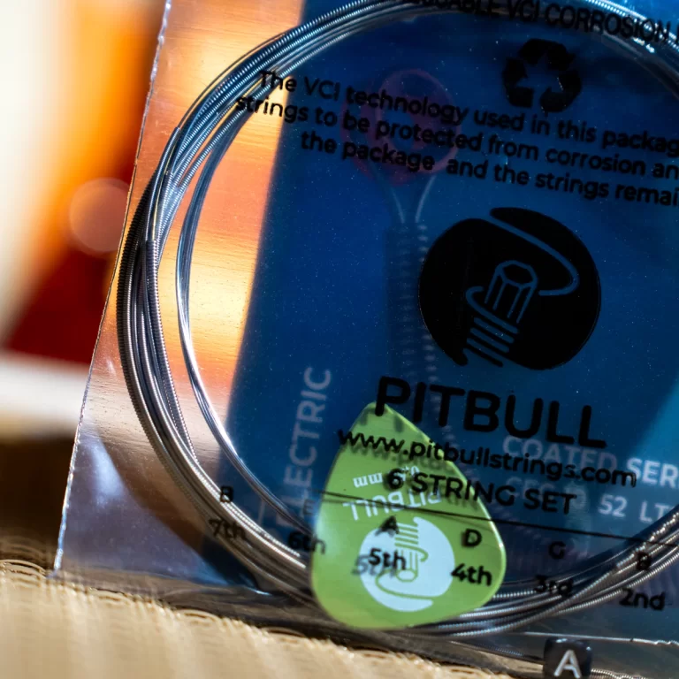 Close-up of a blue Pitbull guitar string set packaging with coiled strings and a green guitar pick resting on it ขายราคาพิเศษ
