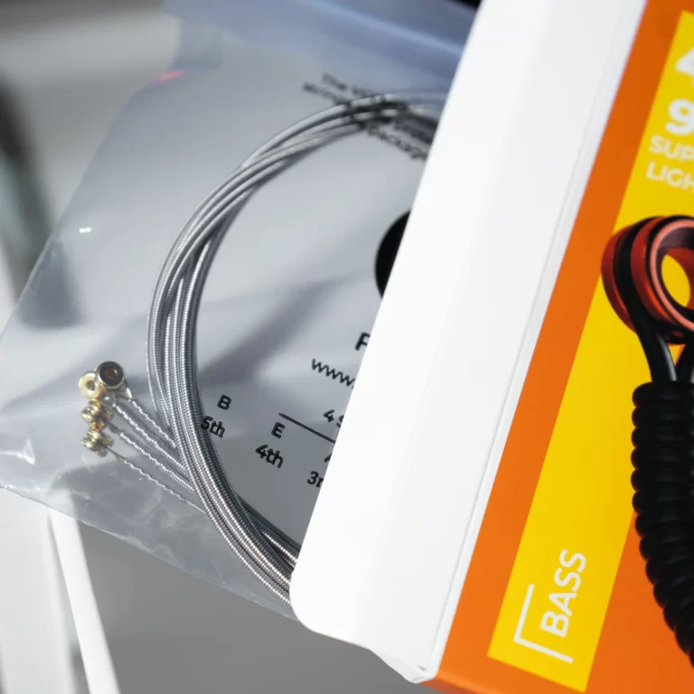 Coiled bass guitar strings with ball-end anchors in a clear plastic bag next to an orange bass packaging box. ขายราคาพิเศษ