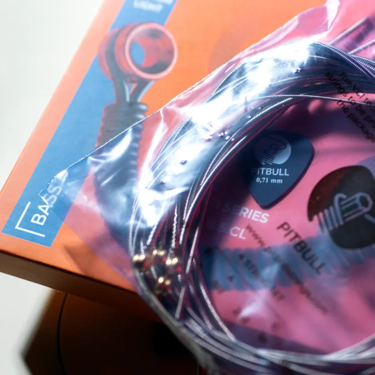 Coiled guitar strings in pink packaging labeled Pitbull, 0.71 mm gauge, resting on an orange box with a blue label ขายราคาพิเศษ