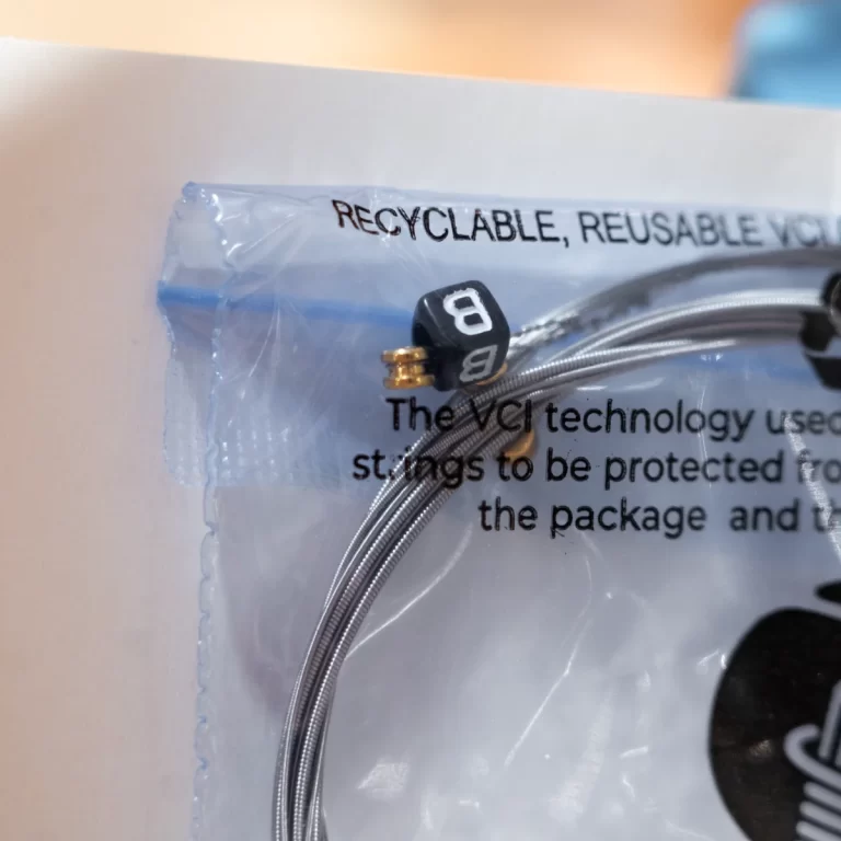 Close-up of a silver braided cable with a small black cube marked '8' and a gold connector, resting on clear packaging that reads 'RECYCLABLE, REUSABLE' and other text. ขายราคาพิเศษ