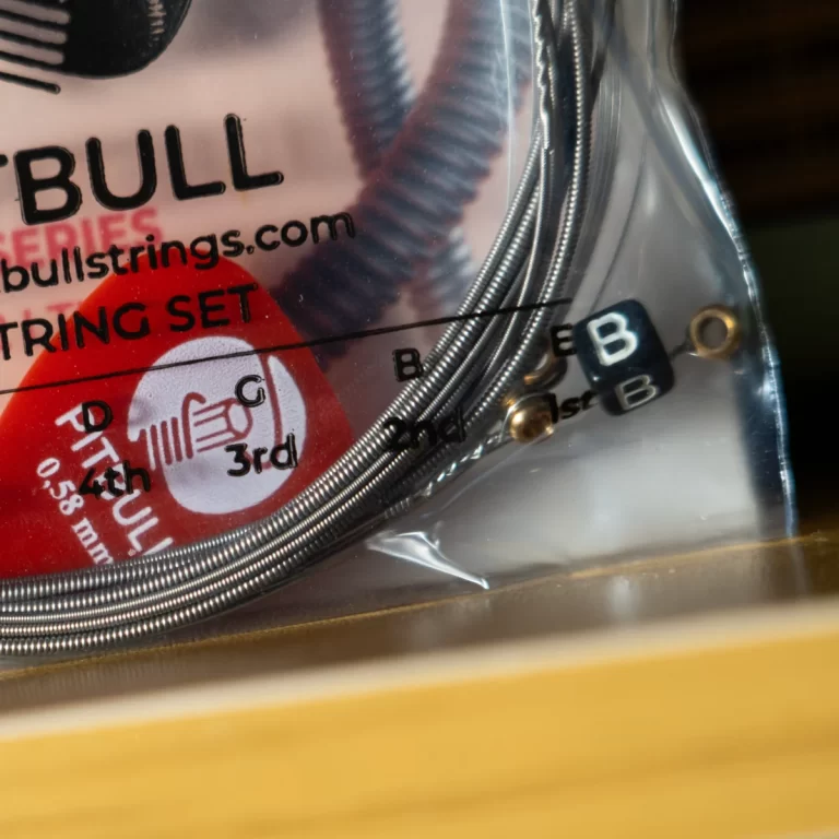 Close-up of a guitar string set package with coiled strings, branding 'BULL' and 'STRING SET' text visible, and small black beads labeled 'B' and '1st'/ 'B' ขายราคาพิเศษ