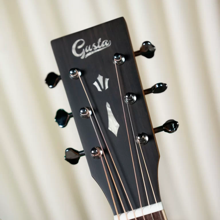 Close-up of a dark guitar headstock with six tuning pegs and strings, showing the brand logo 'Gusta' and a decorative inlay. ขายราคาพิเศษ
