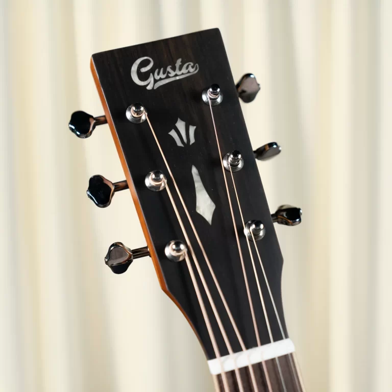 Headstock of a guitar with the Gusta logo and six tuning pegs, close-up view of strings against a light background ขายราคาพิเศษ