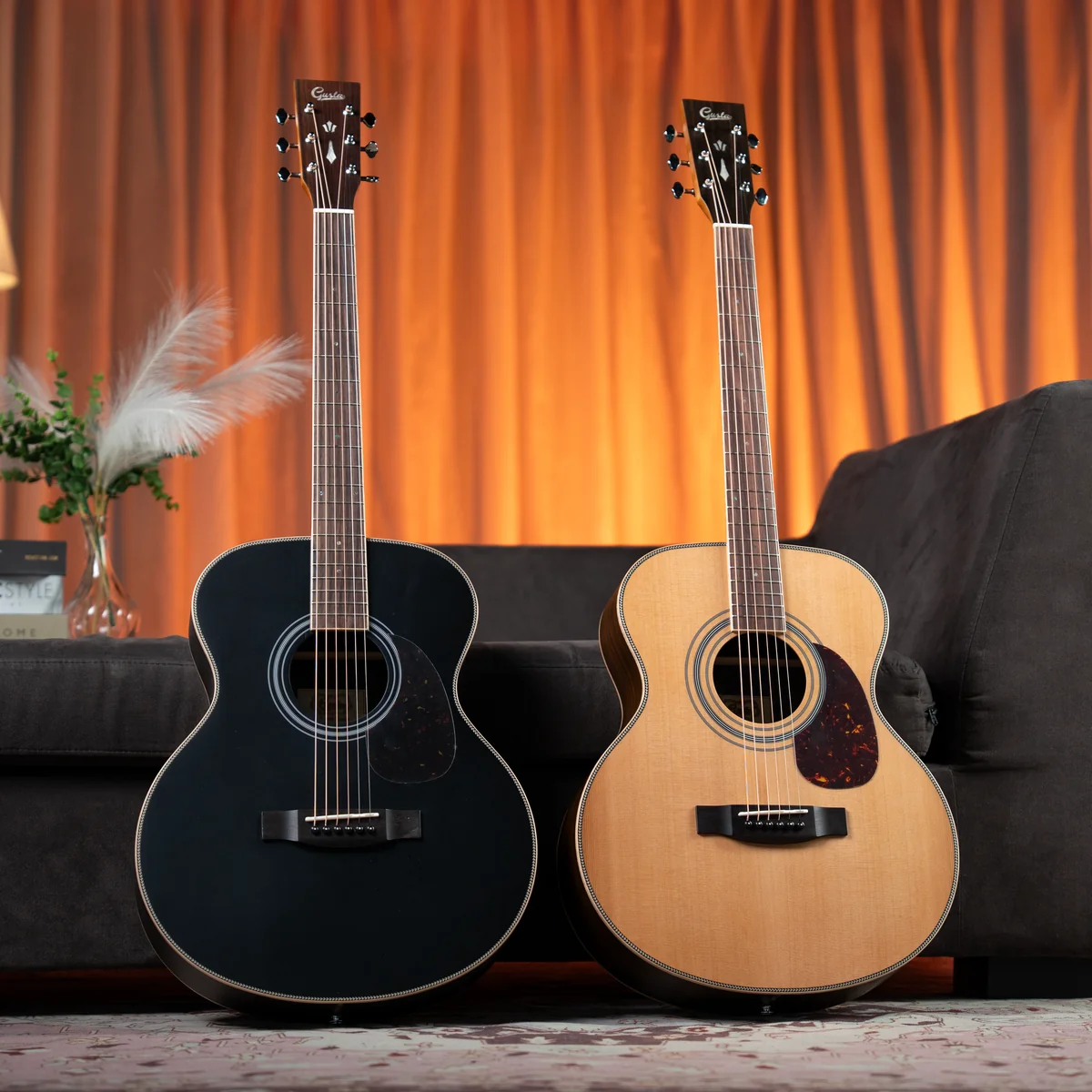 Two acoustic guitars sit on the floor in a living room, one black and one natural wood, with an orange-curtain backdrop and sofa behind them.