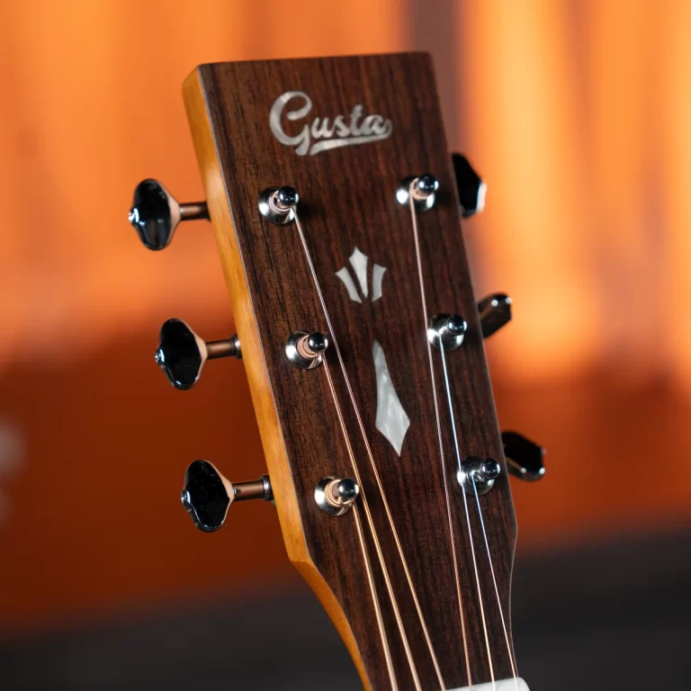 Close-up of a wooden guitar headstock with silver tuning pegs, displaying a Gusta logo and white inlay on a warm orange background. ขายราคาพิเศษ