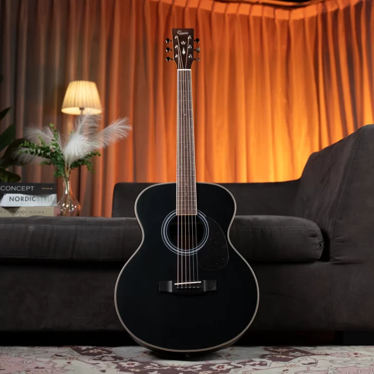 Black acoustic guitar standing upright in a living room with a dark sofa and warm orange curtains behind it. ขายราคาพิเศษ