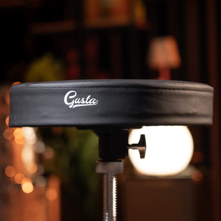 Close-up of a black padded stool seat with the brand name 'Gusta' on a metal adjustable post, blurred warm background. ขายราคาพิเศษ