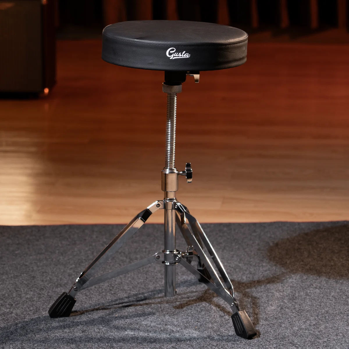Black padded drum throne on a chrome adjustable stand with a three-legged chrome base on a stage floor.