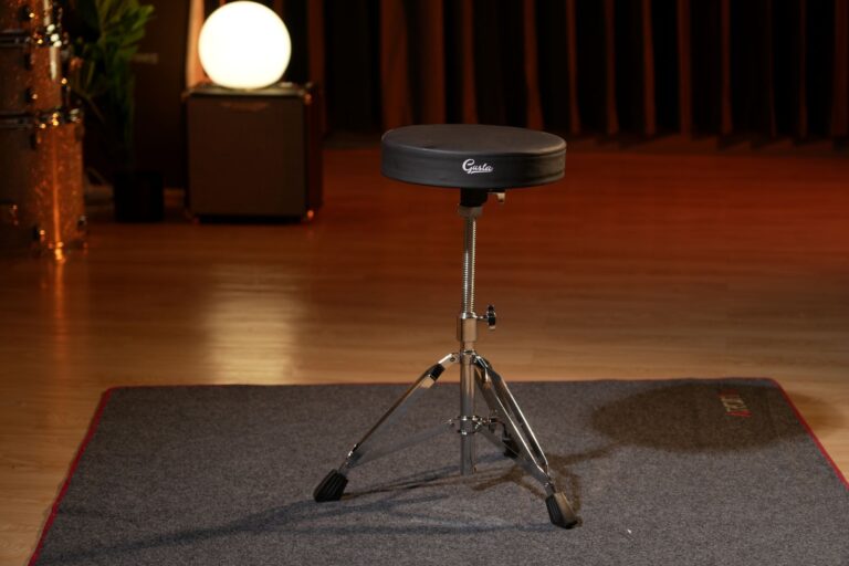 Black round drum throne on a chrome, adjustable stand centered on a warm-lit stage with a wooden floor and red-edge carpet nearby, brand 'Gusta' visible on the seat. ขายราคาพิเศษ