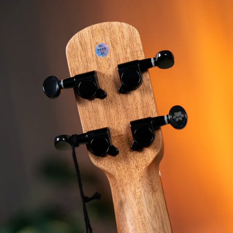 Wooden guitar headstock with four black tuning pegs and a blue QC PASS sticker on the wood, against a warm orange background. ขายราคาพิเศษ