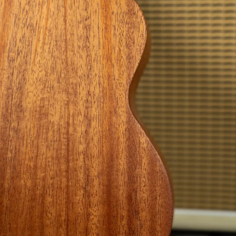 Close-up of a wooden guitar body with visible grain and a curved edge, against a textured amplifier grille in the background. ขายราคาพิเศษ