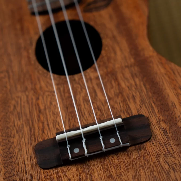 Close-up of a wooden string instrument bridge (ukulele) with a white saddle and four strings crossing over the bridge on a wood-grained body. ขายราคาพิเศษ