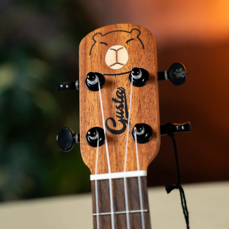 Close-up of a wooden ukulele headstock with four tuning pegs, white strings, and a bear-face carving on the headstock. The brand name is visible in script. ขายราคาพิเศษ