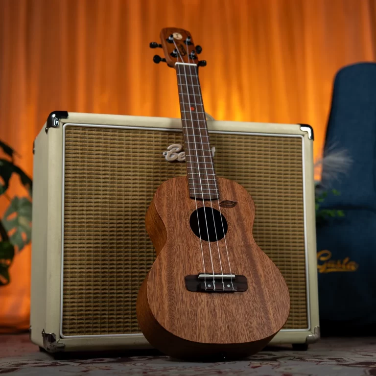 Wooden ukulele leaning against a large beige guitar amplifier with a woven grille and an orange-lit background. ขายราคาพิเศษ