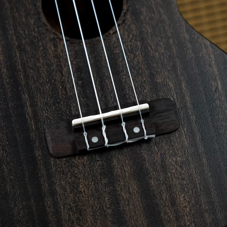Close-up of a dark wood guitar body showing the bridge, white saddle, and four strings anchored at the bridge plate on a textured surface. ขายราคาพิเศษ