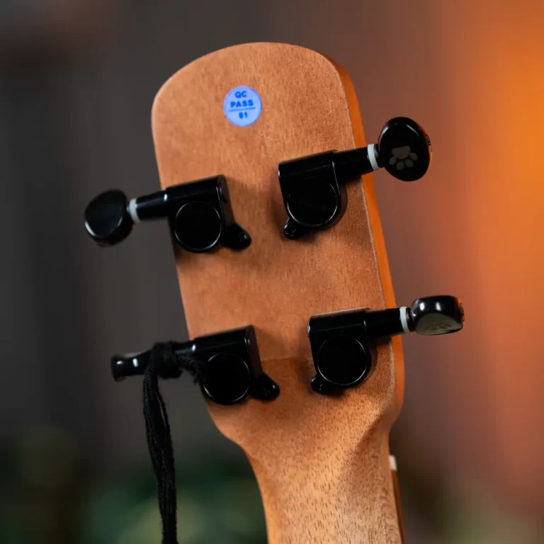 Close-up of a wooden ukulele headstock with four black tuning pegs and a small blue QC PASS sticker on the headstock ขายราคาพิเศษ