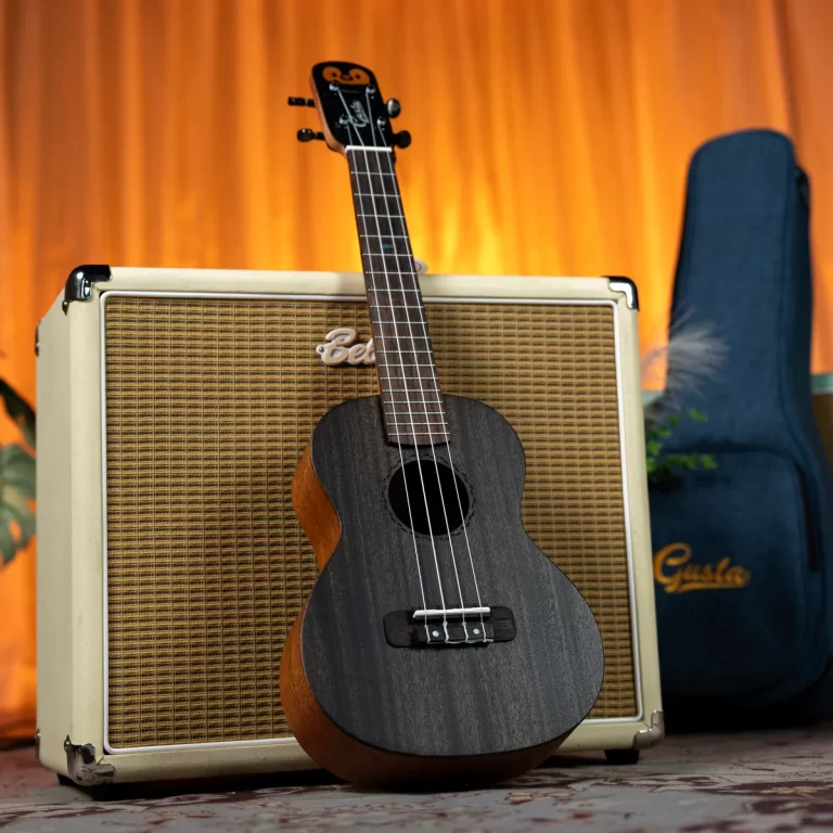 Dark ukulele leaning on a beige woven-grille amplifier with an orange backdrop and a blue guitar case nearby. Par is a musical setup scene. ขายราคาพิเศษ