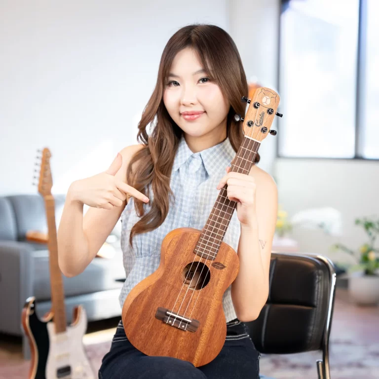 Smiling woman wearing a checkered sleeveless top holds a wooden ukulele and points to it with her right hand in a bright, cozy room. ขายราคาพิเศษ