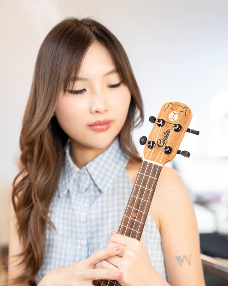 Young woman with long brown hair in a light blue checkered sleeveless top, holding a wooden ukulele and looking down at the fretboard. ขายราคาพิเศษ