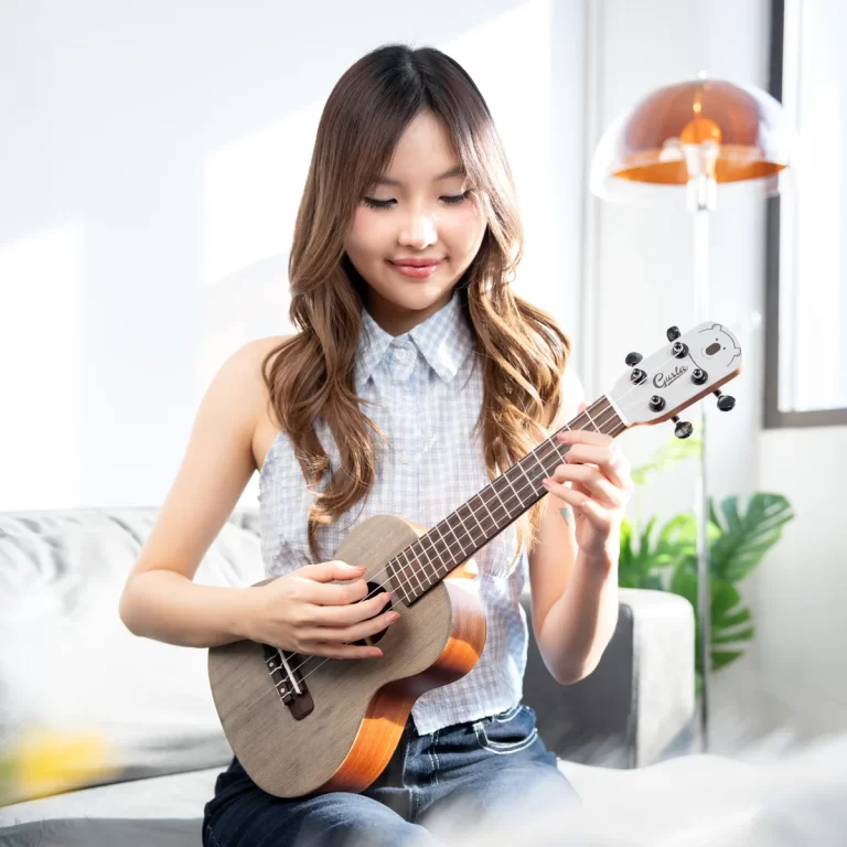 Young woman sits on a sofa, focused on playing a small ukulele in a bright, cozy living room. ขายราคาพิเศษ