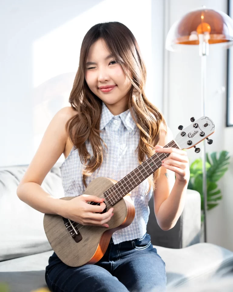 Young woman with wavy hair winking while playing a ukulele indoors on a sofa. ขายราคาพิเศษ