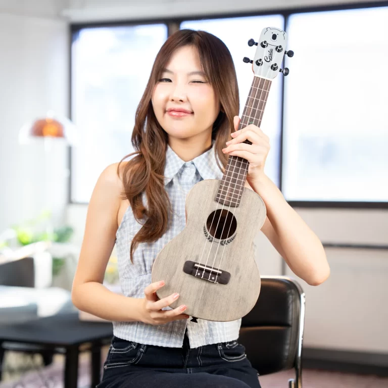 Smiling woman sits indoors, holding a small gray ukulele. ขายราคาพิเศษ