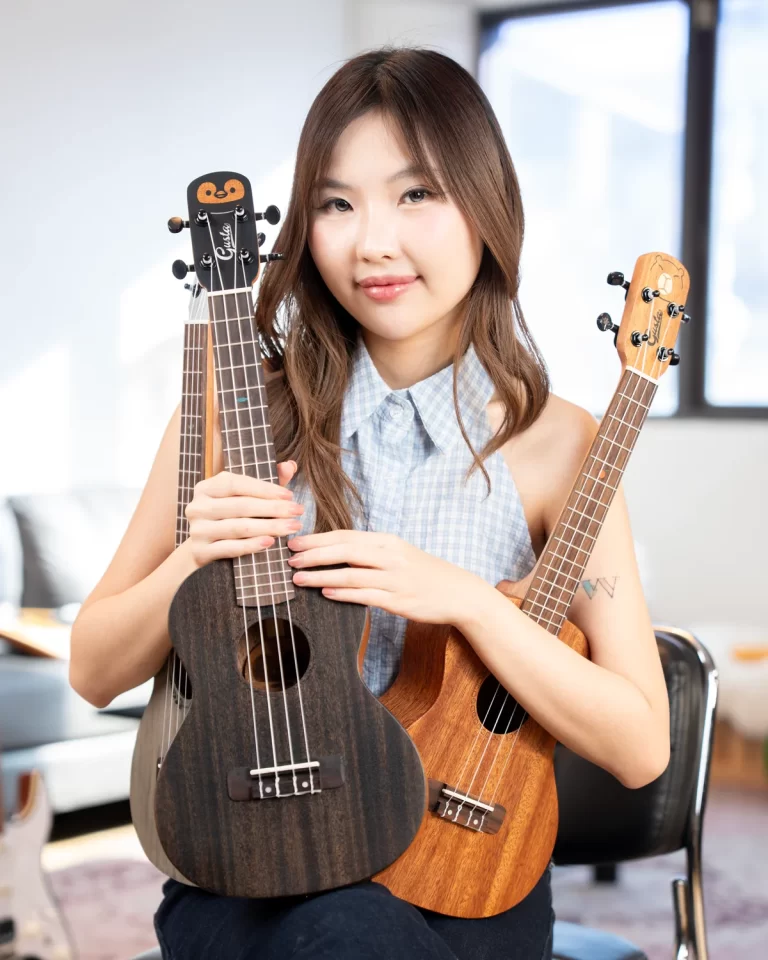 Young woman sits indoors, smiling, holding two ukuleles—one dark wood and one lighter wood—up to the camera. ขายราคาพิเศษ