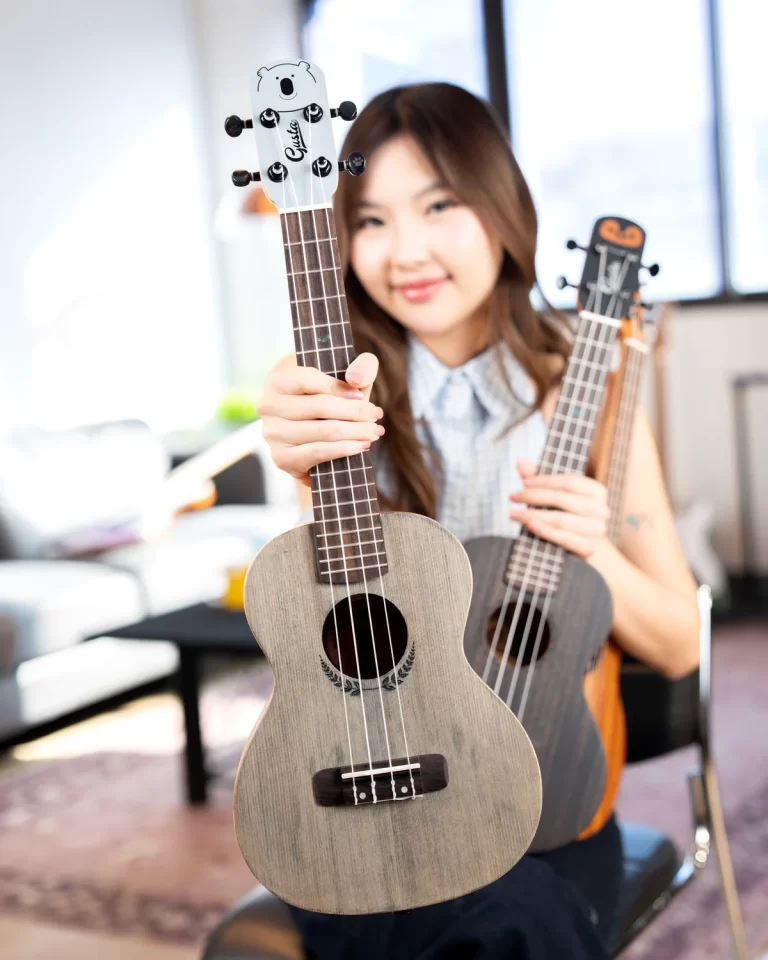 Smiling woman in a bright room holds a light wood ukulele close to the camera, two ukuleles visible in her hands. ขายราคาพิเศษ