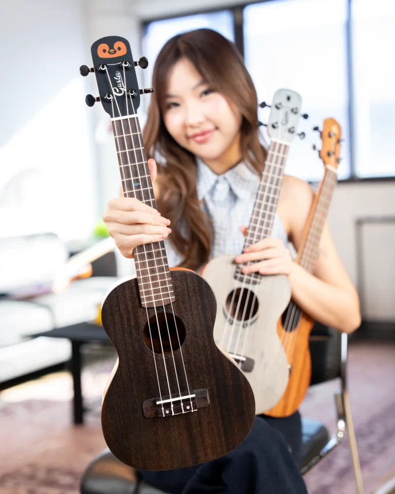 Woman indoors holding up a dark wood ukulele toward the camera, with two other ukuleles nearby and a smile on her face. ขายราคาพิเศษ