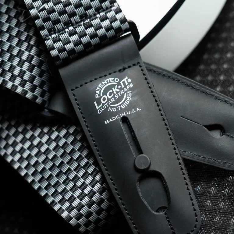 Close-up of a black leather guitar strap with the 'Lock-It' patent label and a slot for attaching to the guitar, over a woven textured background. ขายราคาพิเศษ