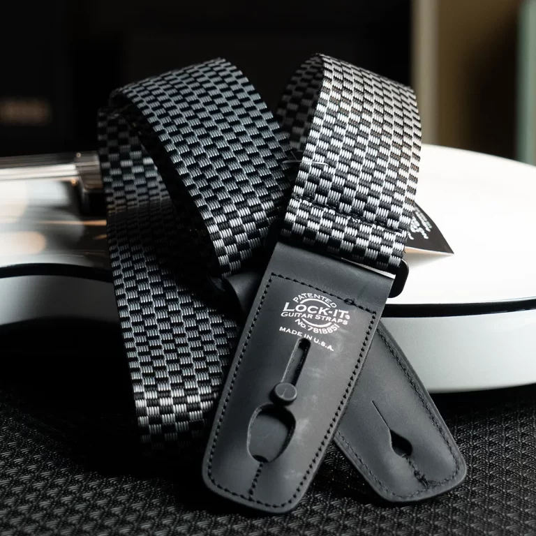 Checkerboard black-and-gray guitar strap with leather ends draped over a white guitar body, close-up detail of the webbing pattern and hardware. ขายราคาพิเศษ