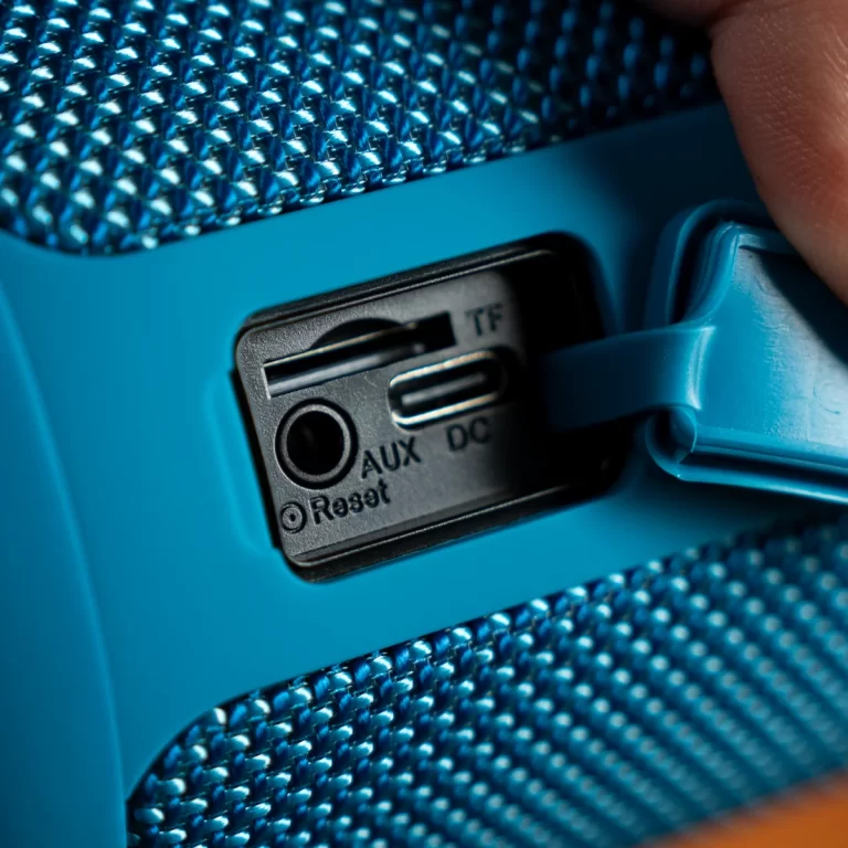 Close-up of a blue portable speaker's control panel showing AUX and DC ports with a TF card slot and a finger lifting a latch. ขายราคาพิเศษ