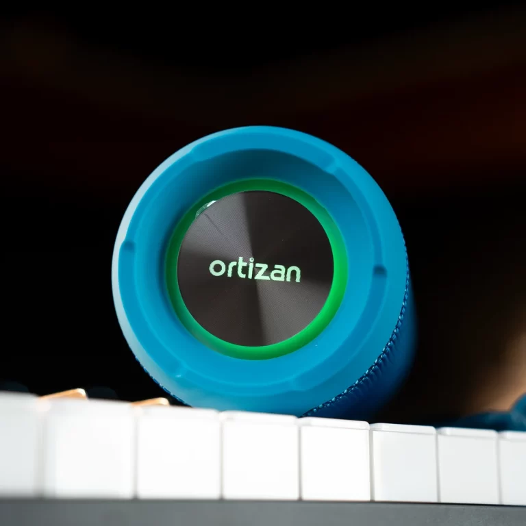 Blue over-ear headphone earcup resting on piano keys, showing the Ortizan logo with a green ring around it (informative product shot). ขายราคาพิเศษ