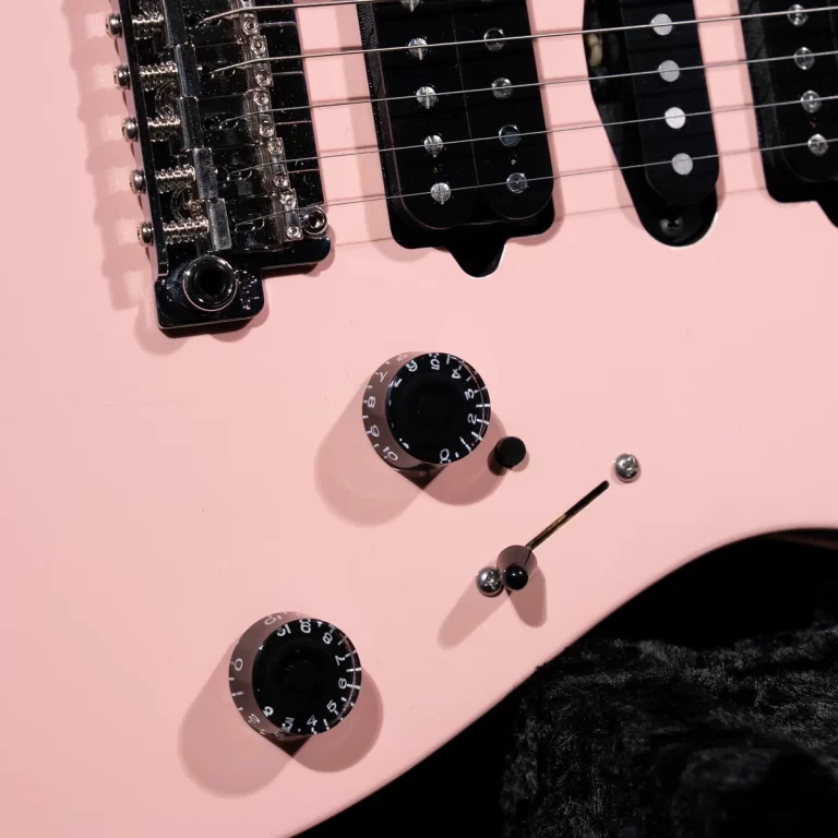 Close-up of a pink electric guitar body with chrome hardware, showing a tremolo bridge, three black pickups, and two large black control knobs with white numbers. ขายราคาพิเศษ