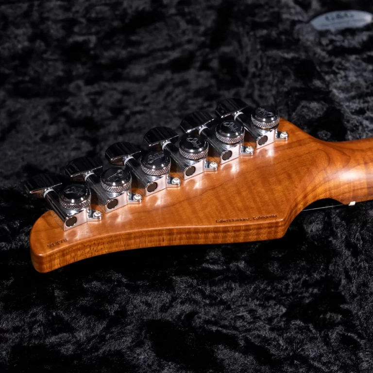 Close-up of a wooden guitar headstock with chrome tuning pegs, resting on a dark plush surface; along the edge there is a faint engraving ending with 'California'. ขายราคาพิเศษ