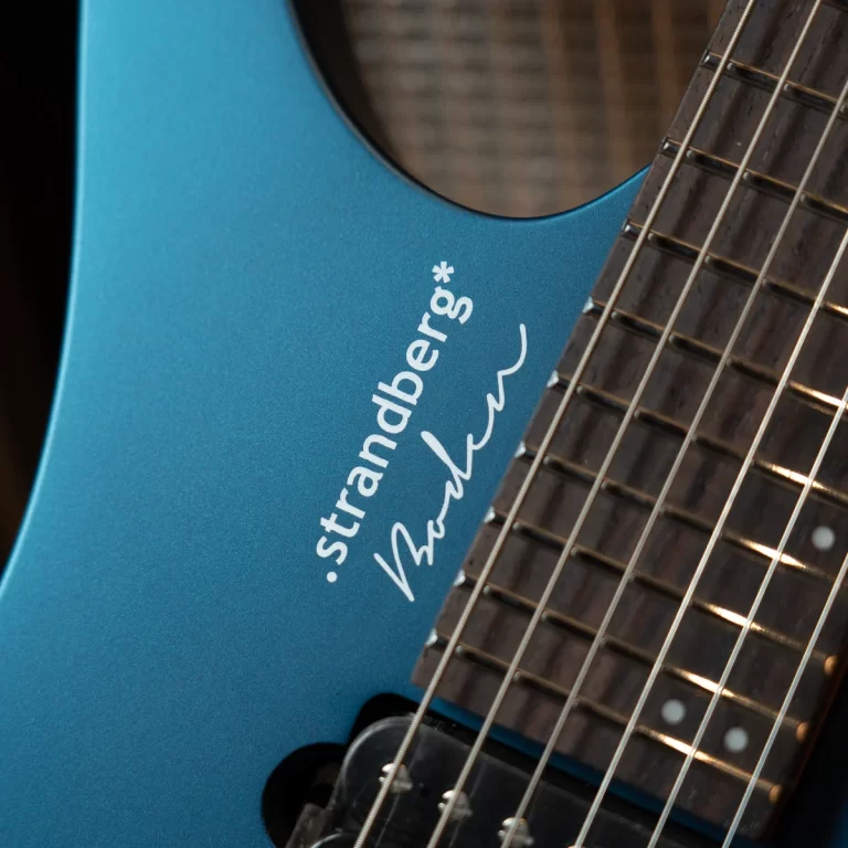 Close-up of a blue Strandberg electric guitar body with a partial dark fretboard and strings; the brand name and a handwritten signature are visible on the body. ขายราคาพิเศษ
