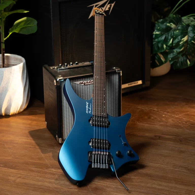 Blue Strandberg electric guitar leaning against a black guitar amplifier on a wooden floor, with potted plants in the background. ขายราคาพิเศษ